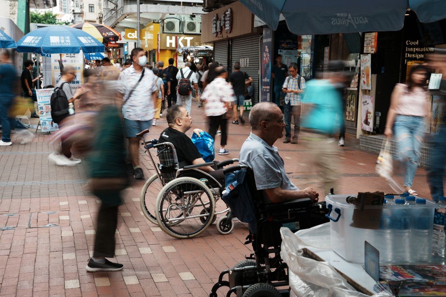 Pedestres, incluindo pessoas em cadeiras de rodas, circulam por uma rua urbana movimentada.