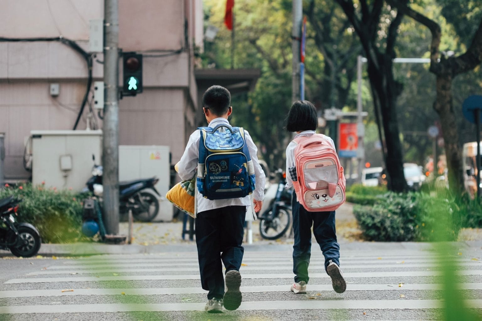 Duas crianças com mochilas atravessam uma rua pela faixa de pedestres.