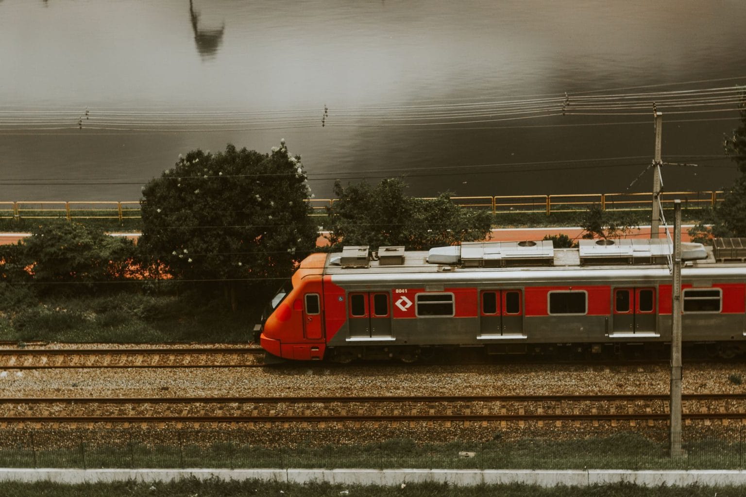 Trem vermelho circula por via férrea urbana às margens de um rio, em São Paulo.